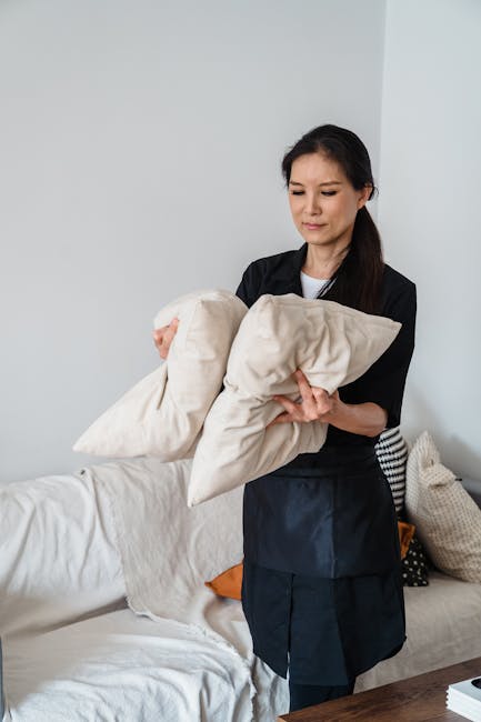 A professional cleaner dressed in white protective clothing, including a hood, mask, and blue gloves, is conducting surface cleaning and sanitisation on a dark brown upholstered armchair in a modern l