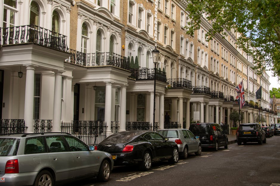 Image of a row of elegant, white Victorian-style terraced houses with ornate architectural details, balconies, and large windows, situated along a busy street with parked cars and black street lamps. 