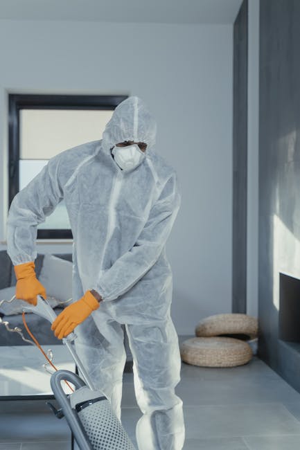 A professional cleaner from Belgravia Cleaner, dressed in white protective clothing, gloves, and blue shoe covers, is performing surface cleaning with a vacuum and hose attachment on a plush, beige ca